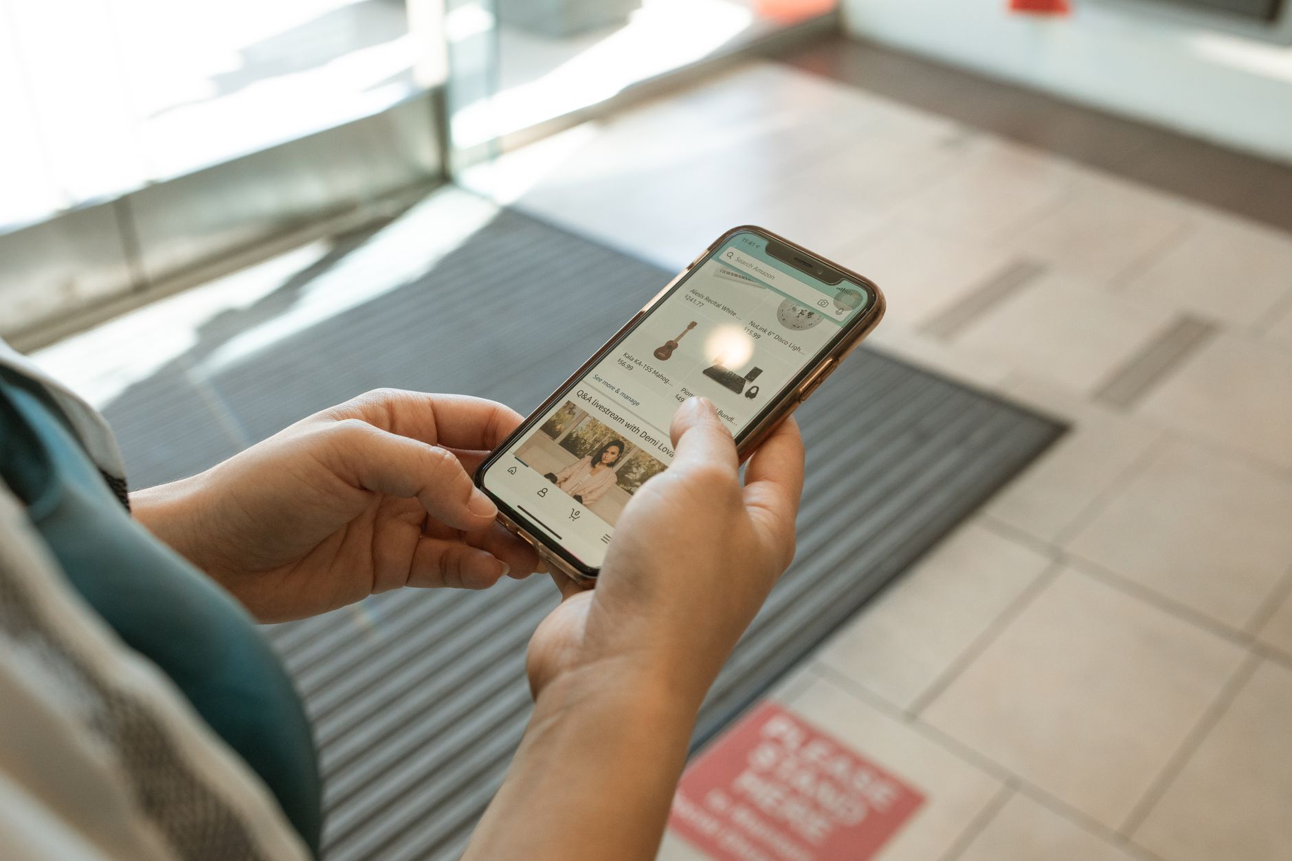 Deux mains tiennent un smartphone affichant une application de boutique en ligne, dans un hall au sol carrelé avec un tapis rayé.