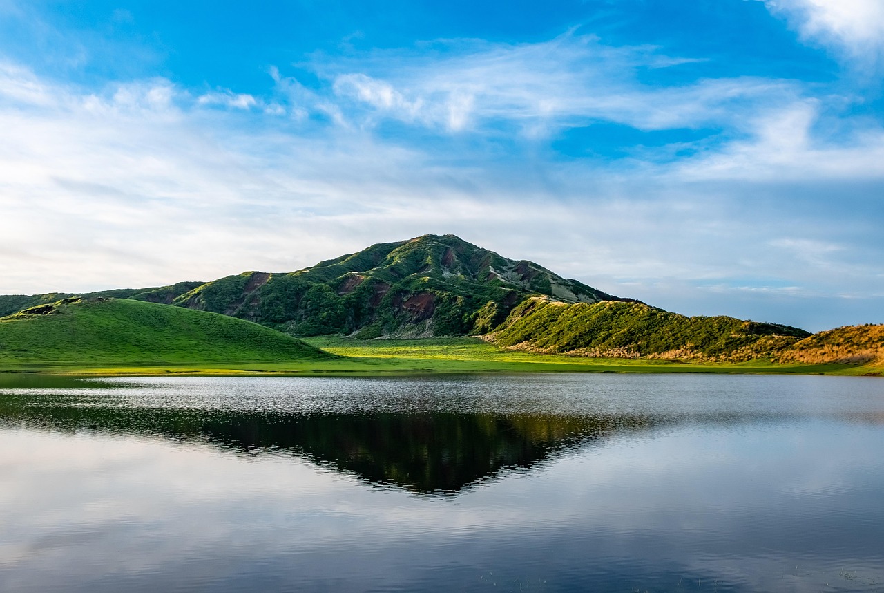 Lac calme dont la surface reflète une colline verte, sous un ciel bleu traversé de nuages