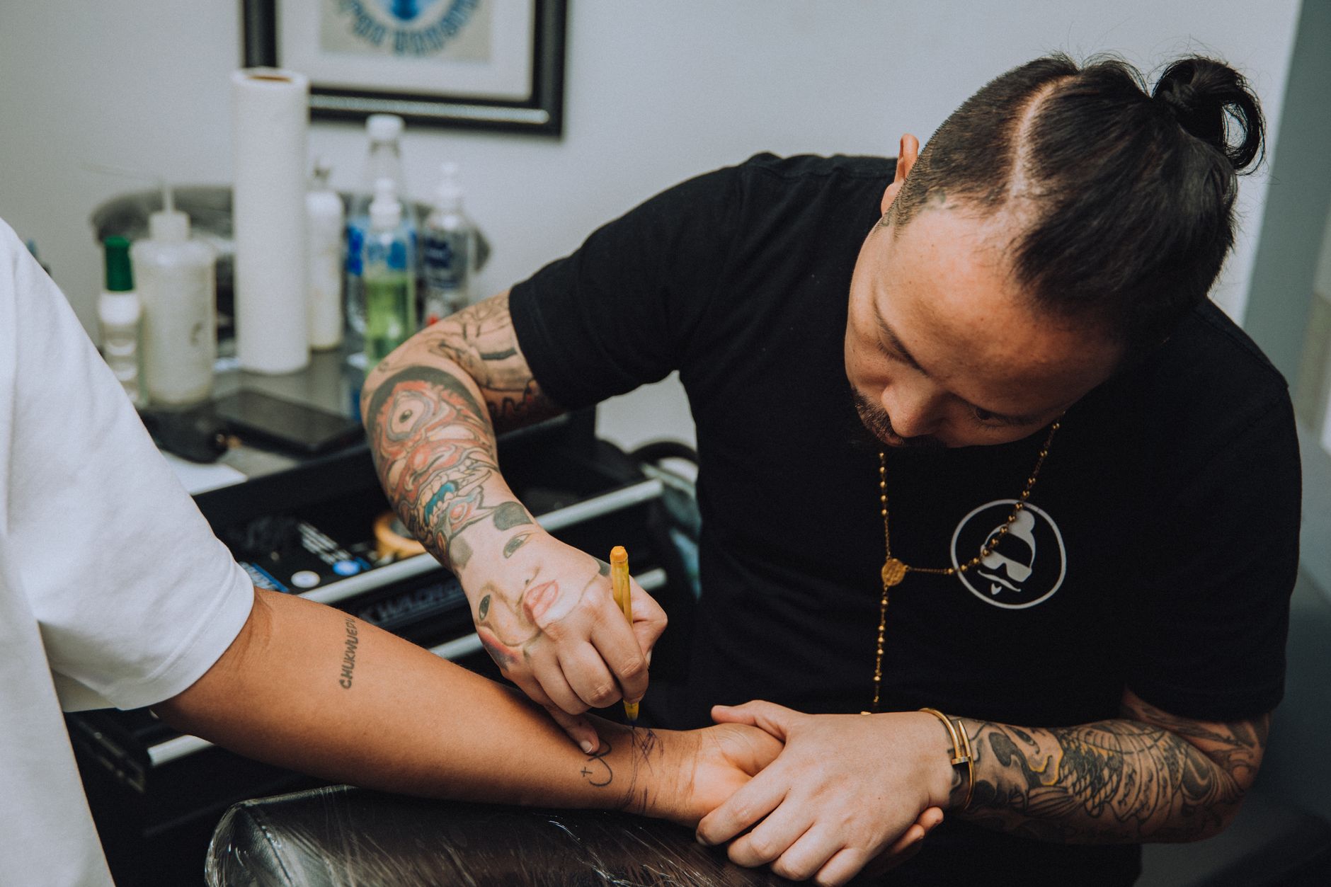 Un tatoueur aux bras tatoués dessine au feutre un motif sur l’avant-bras d’un client, dans un salon avec flacons et matériel en arrière-plan.